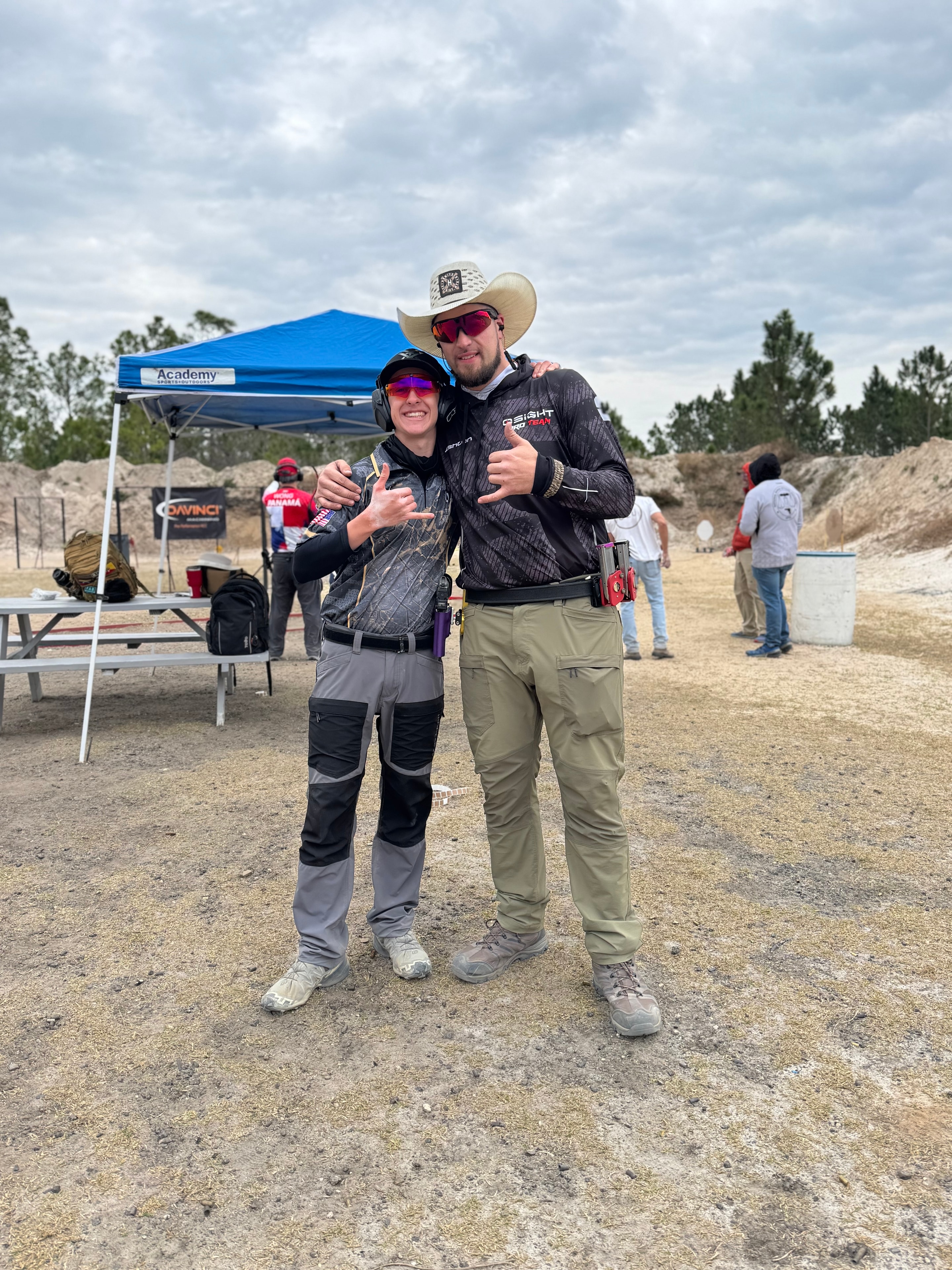 Two competitors posing together at an outdoor USPSA match with an Academy Sports tent in the background