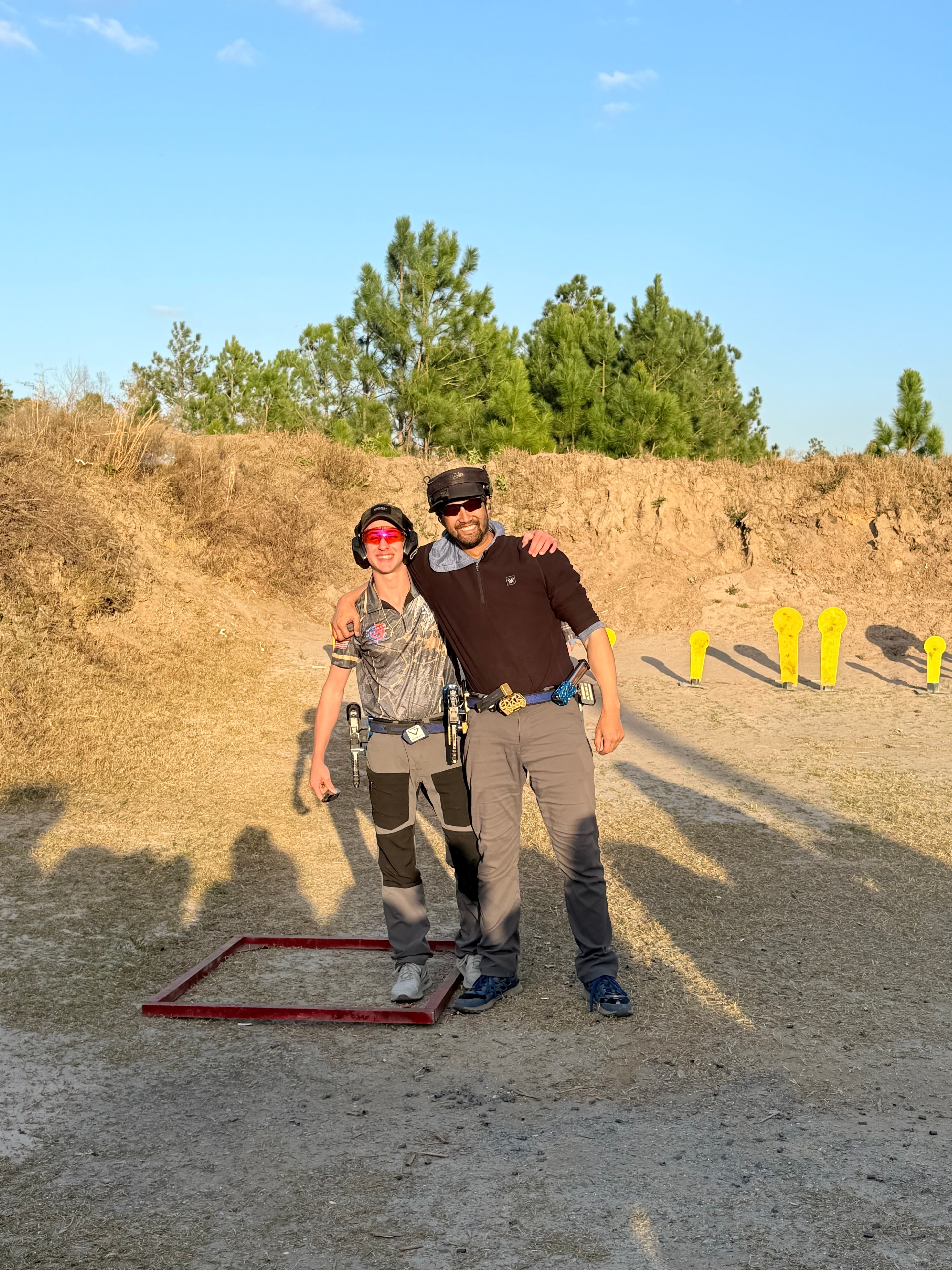 Two competitors at an outdoor shooting range with yellow steel targets in the background