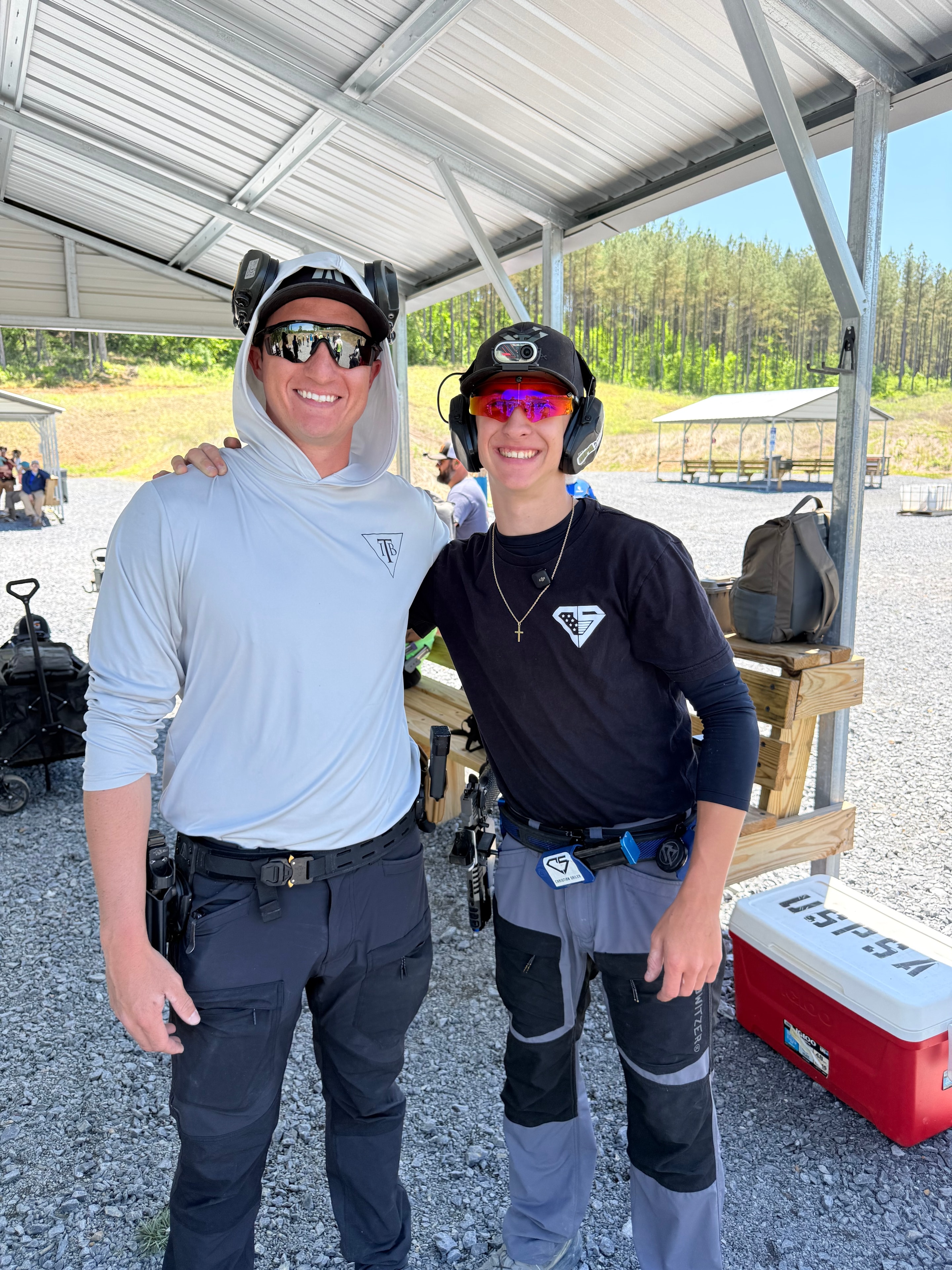 Two USPSA competitors smiling together at a shooting range bay, both in full competition gear
