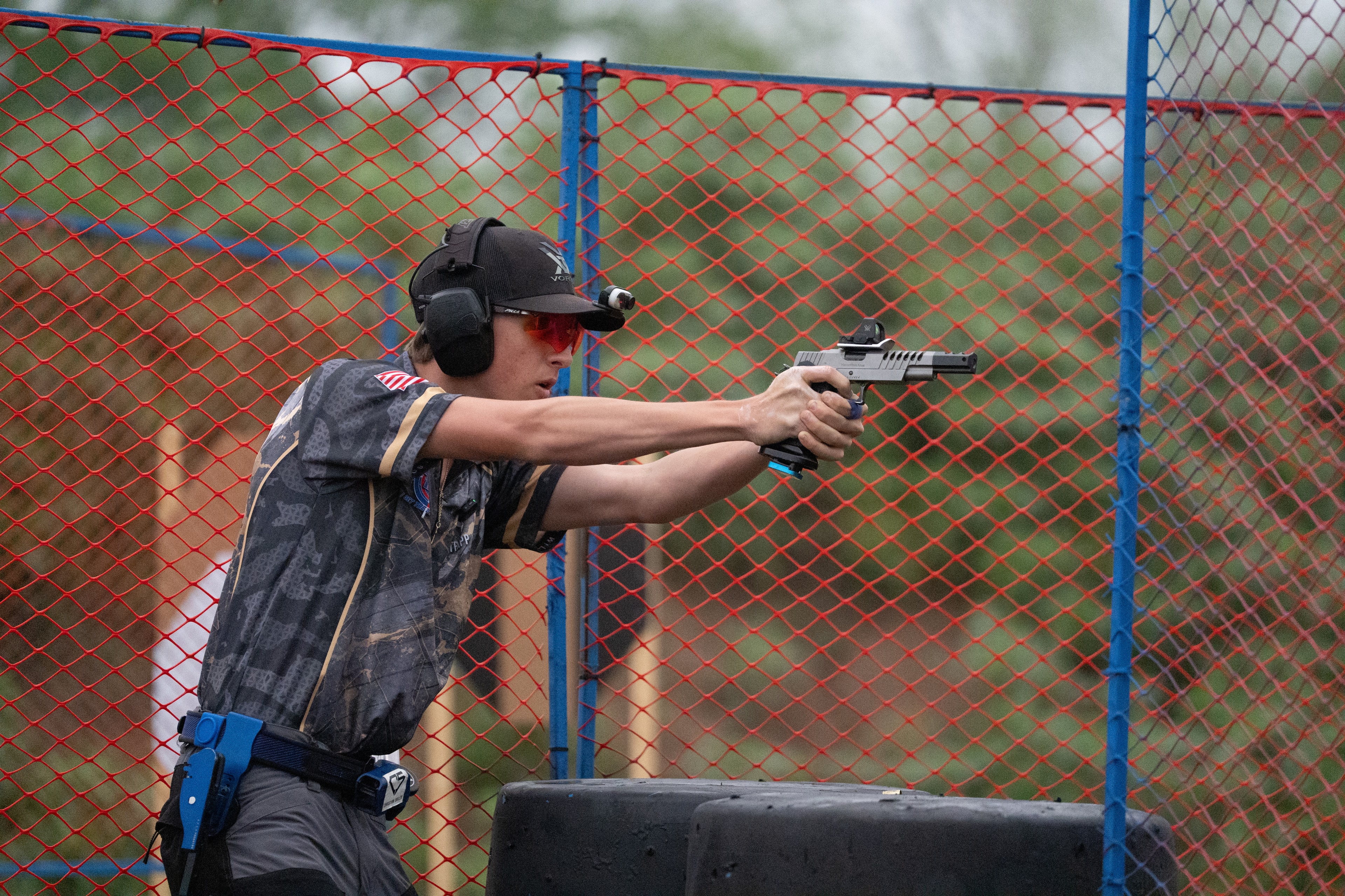 Tripp Wilson aiming a pistol at a USPSA competition stage with a red chain-link fence backdrop