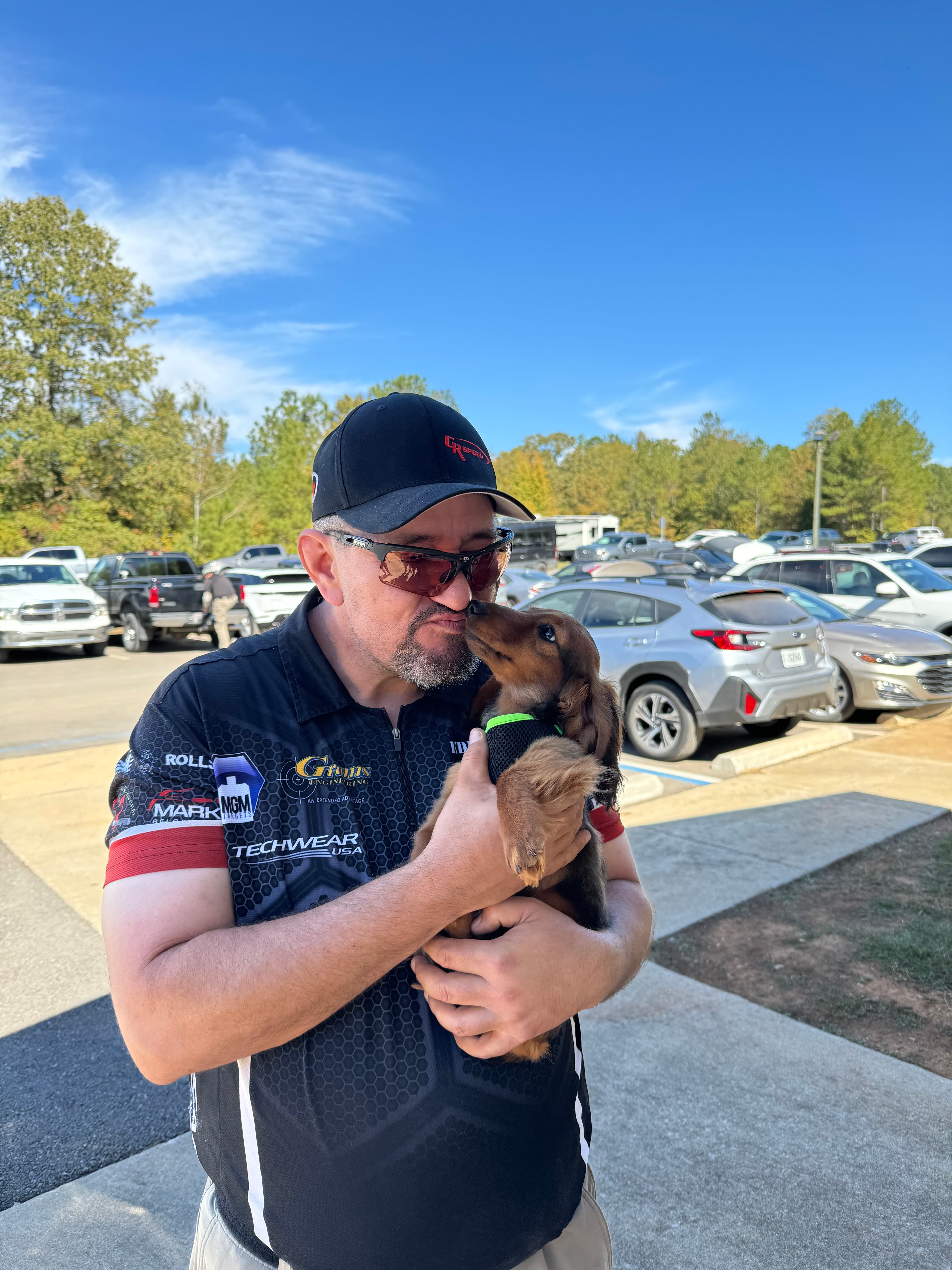 Tripp Wilson holding a puppy at a shooting match, wearing High Standards Shooting Solutions competition gear